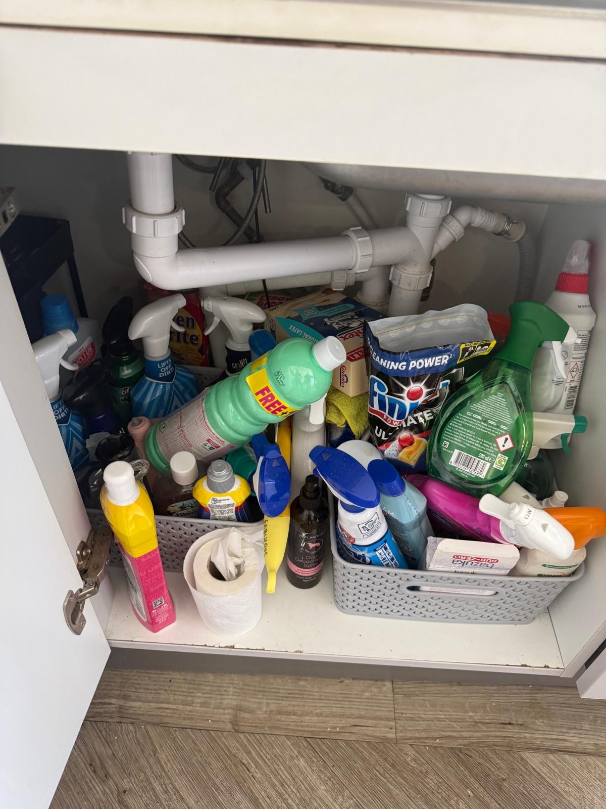 Cluttered under-sink cupboard filled with cleaning sprays before a kitchen organisation session