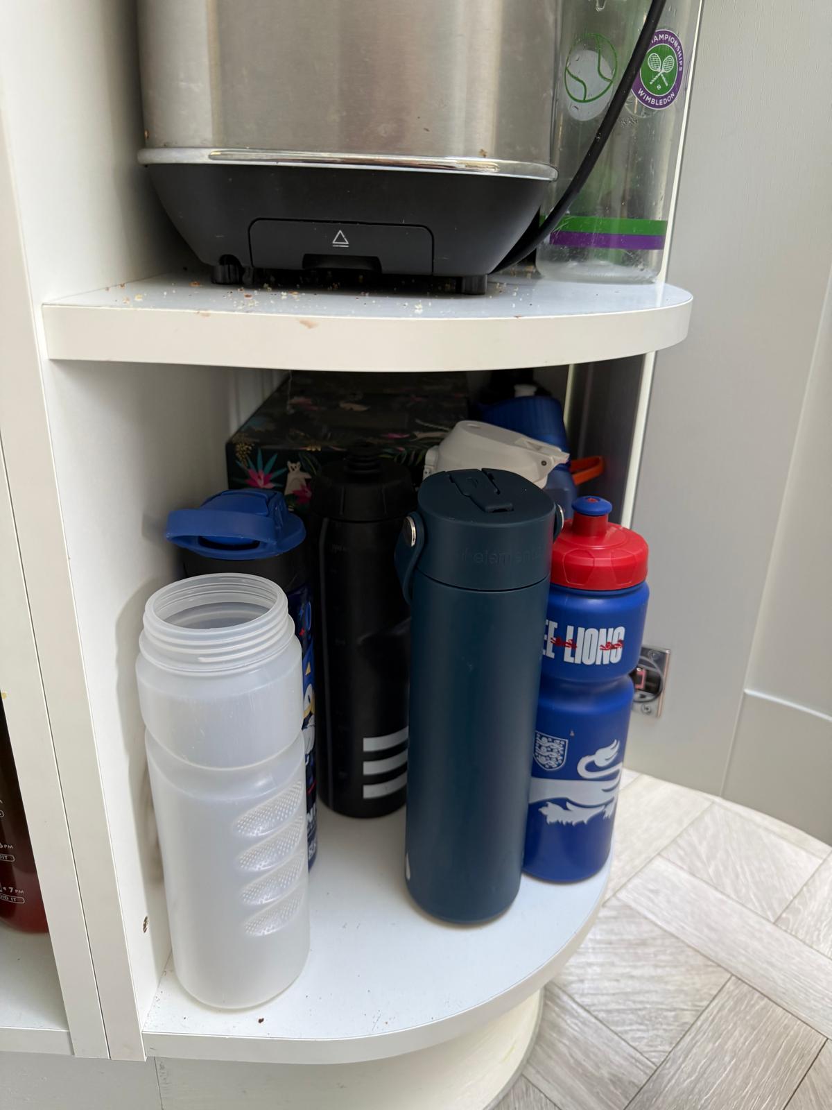 Water bottles and kettle shelf inside a kitchen unit before organisation
